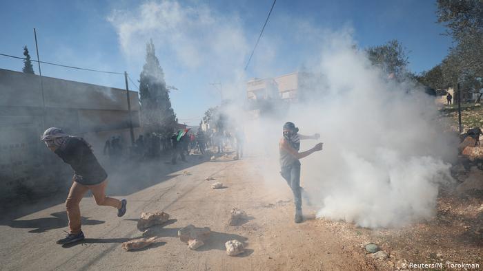 Палестинцы в масках бегут в дыму по дороге, под ногами у них валяются камни